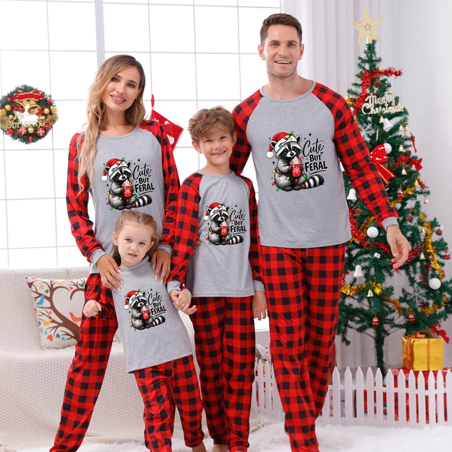 A family of four wearing matching gray and red buffalo plaid pajamas with a raccoon graphic that says 'Cute But Feral' posed in front of a decorated Christmas tree and holiday wreath.