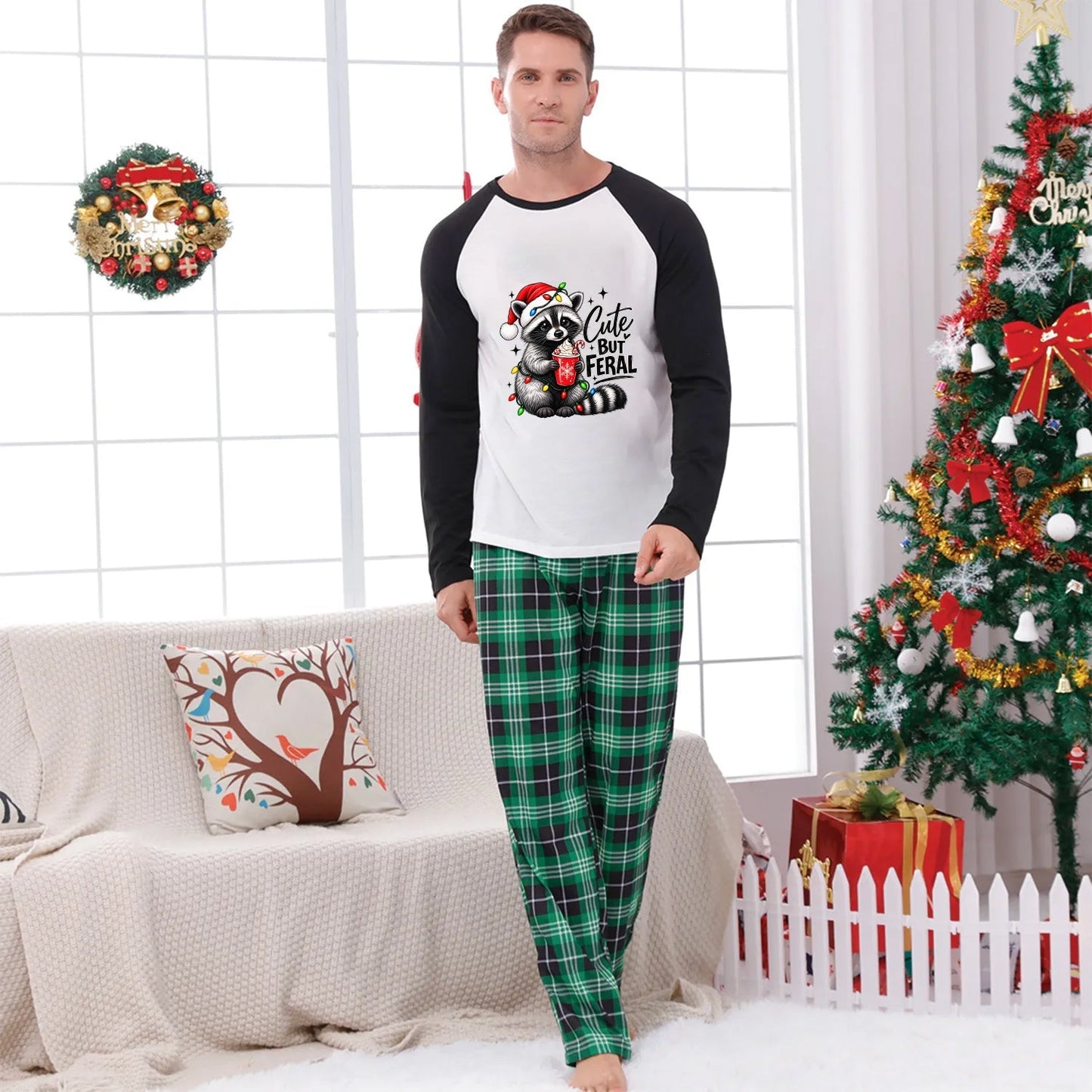 A man wearing a Christmas-themed pajama set stands in a decorated living room. The set includes a white and black raglan long-sleeve shirt with a raccoon graphic and the text ' Cute but Feral' on the front, paired with green plaid pajama pants.