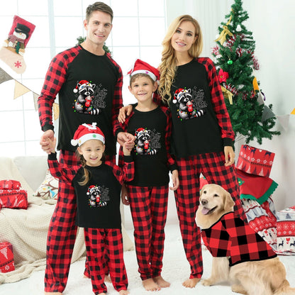 A family of four wearing matching black and red plaid Christmas pajamas with a raccoon graphic, standing in a decorated living room with a dog wearing a matching pajama set. The pajamas feature a black body with red and black buffalo plaid sleeves and legs, and the front has a graphic of a raccoon holding a Christmas tree with the text 'Cute but Feral'.