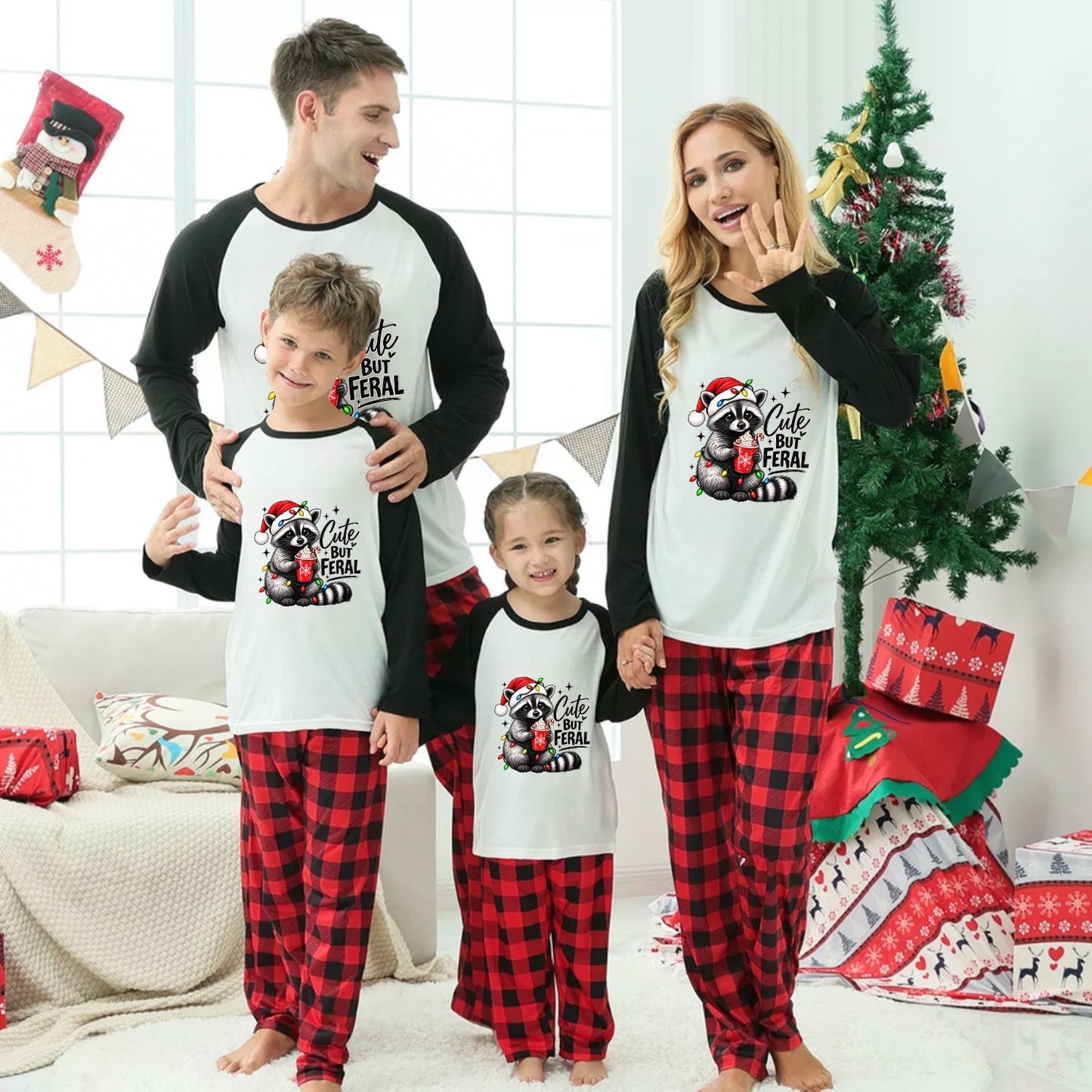 A family of four wearing matching Christmas pajamas stands together in a decorated living room. The pajamas feature a white front with black sleeves and a black-and-red raccoon graphic with the text 'Cute but Feral,' paired with red-and-black plaid pants.