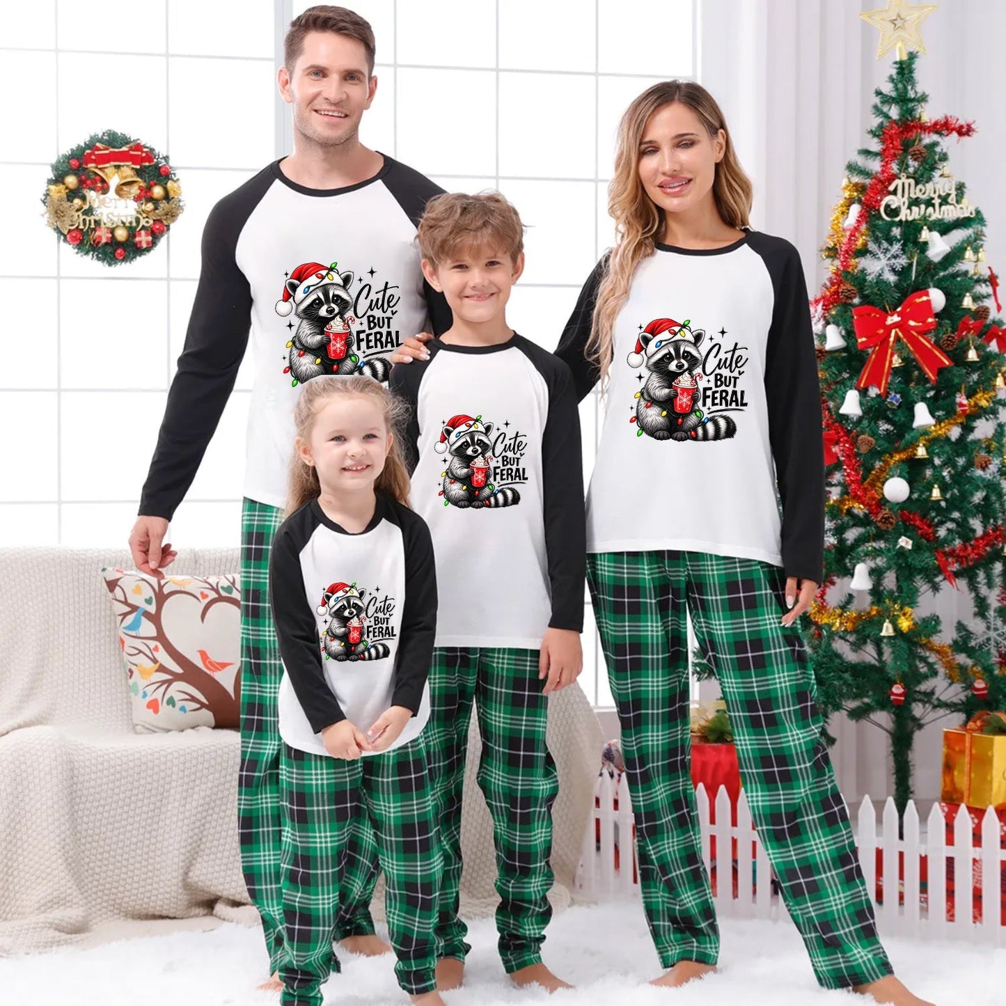 A family of four wearing matching Christmas raglan pajamas stands together in a decorated living room with a Christmas tree, wreath, and couch. The pajamas feature a white front with black sleeves and a black-and-red raccoon graphic with the text 'Cute But Feral' on the chest, paired with green plaid pants.