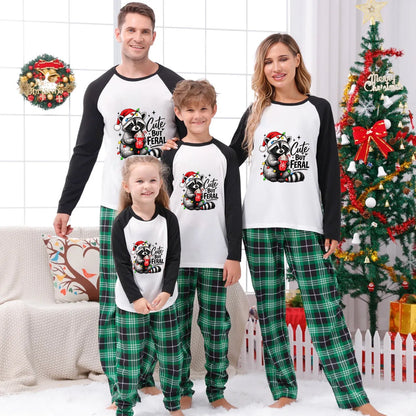 A family of four wearing matching Christmas raglan pajamas stands together in a decorated living room with a Christmas tree, wreath, and couch. The pajamas feature a white front with black sleeves and a black-and-red raccoon graphic with the text 'Cute But Feral' on the chest, paired with green plaid pants.