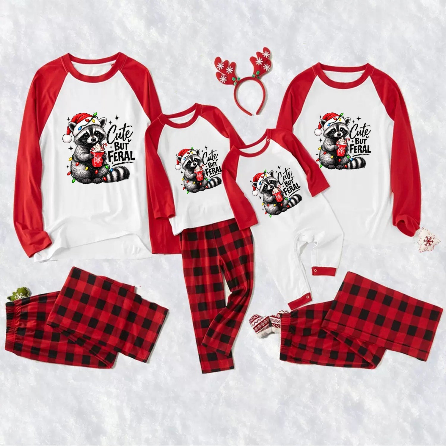 Three matching family Christmas pajama sets laid out on a white textured background, including long-sleeve raglan tops with red sleeves and a raccoon graphic that reads 'Cute But Feral,' red-and-black plaid pants, and a red reindeer antler headband.