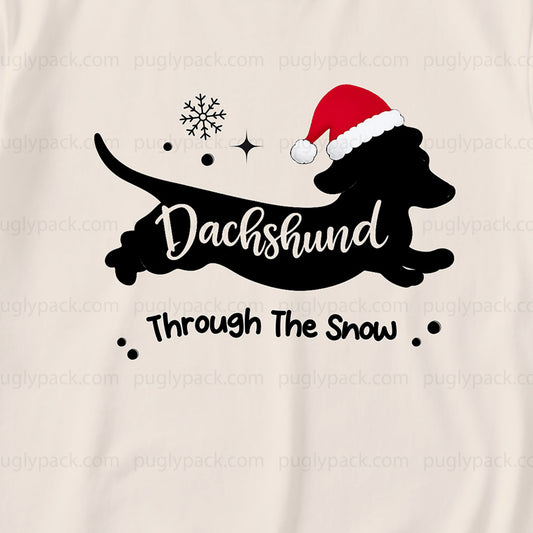 A close-up of a Cream-coloRed garment featuring a Black graphic of a dachshund wearing a Red Santa hat, with the text 'Dachshund Through The Snow' and small snowflake and star accents.