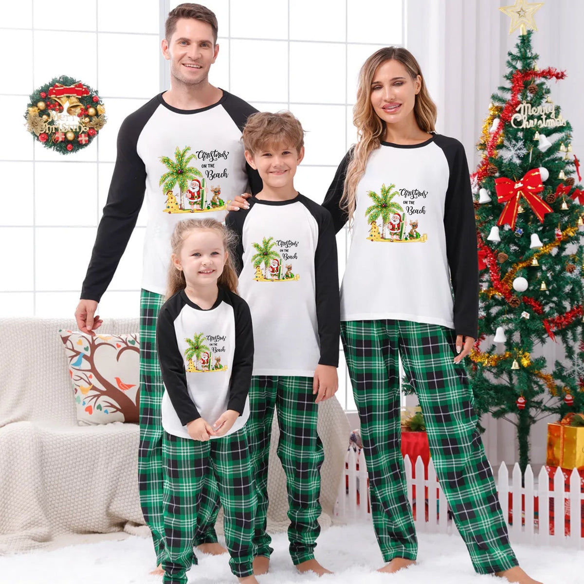 A family of four wearing matching Christmas-themed raglan pajamas stands together in a decorated living room. The pajama set has a White front with Black sleeves and a colorful tropical Santa graphic, paiRed with Green plaid pants.