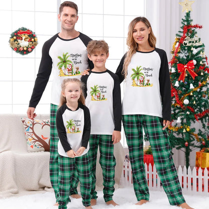 A family of four wearing matching Christmas-themed raglan pajamas stands together in a decorated living room. The pajama set has a White front with Black sleeves and a colorful tropical Santa graphic, paiRed with Green plaid pants.