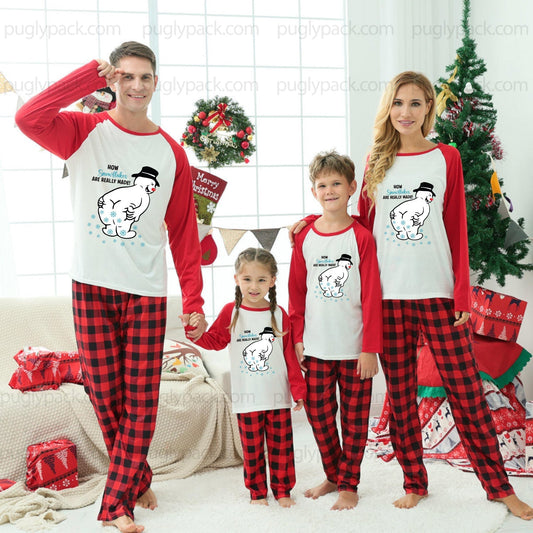 A family of four wearing matching Christmas-themed pajamas, standing together in a decorated living room with a Christmas tree, wreath, and holiday decorations. The pajama set features a White and Red long-sleeve top with a cartoon snowman graphic and Red-and-Black buffalo plaid pants.