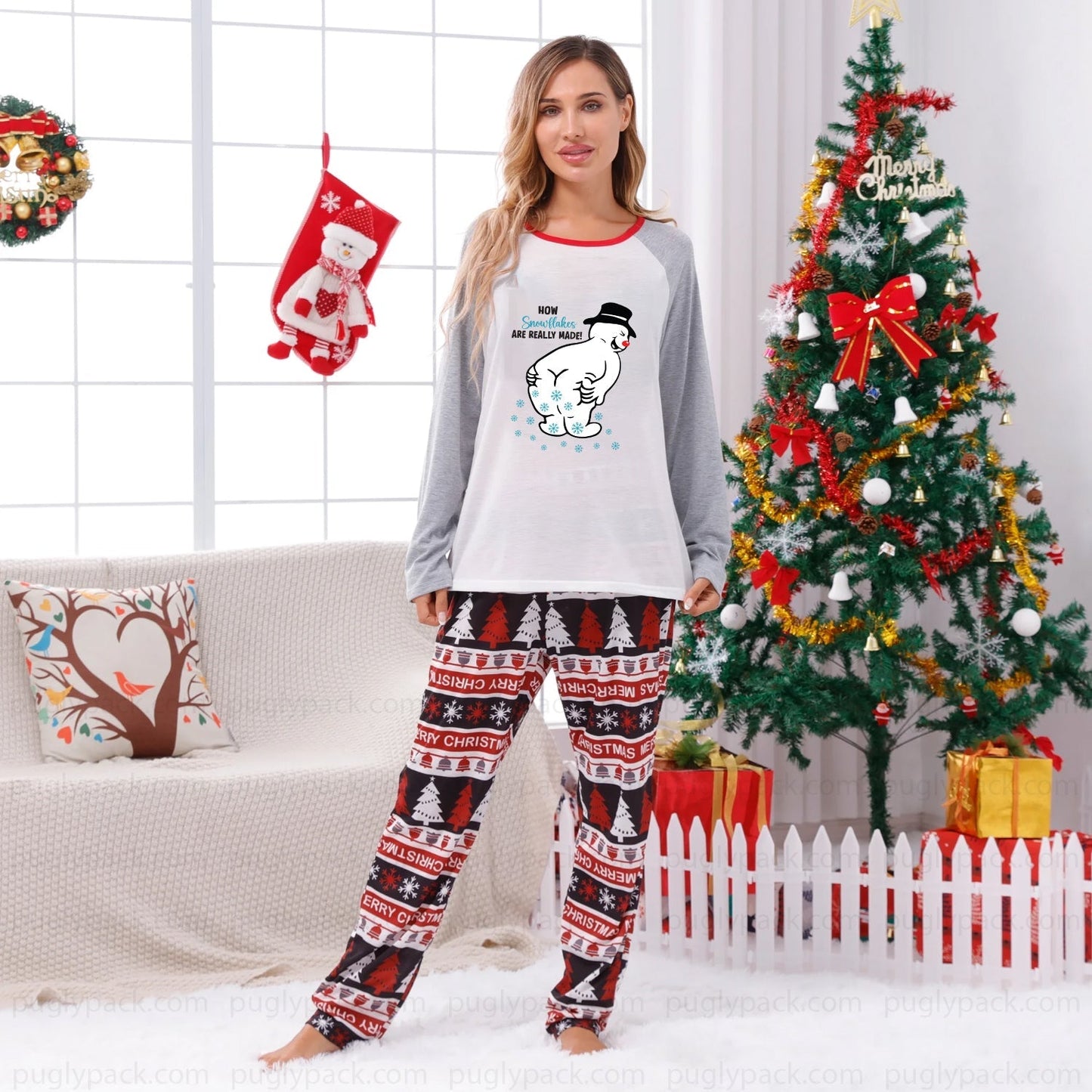 A woman modeling a two-piece Christmas pajama set in a decorated living room. The set features a White long-sleeve top with Gray sleeves and a cartoon snowman graphic, paiRed with matching patterned pants in Red, Black, and White.