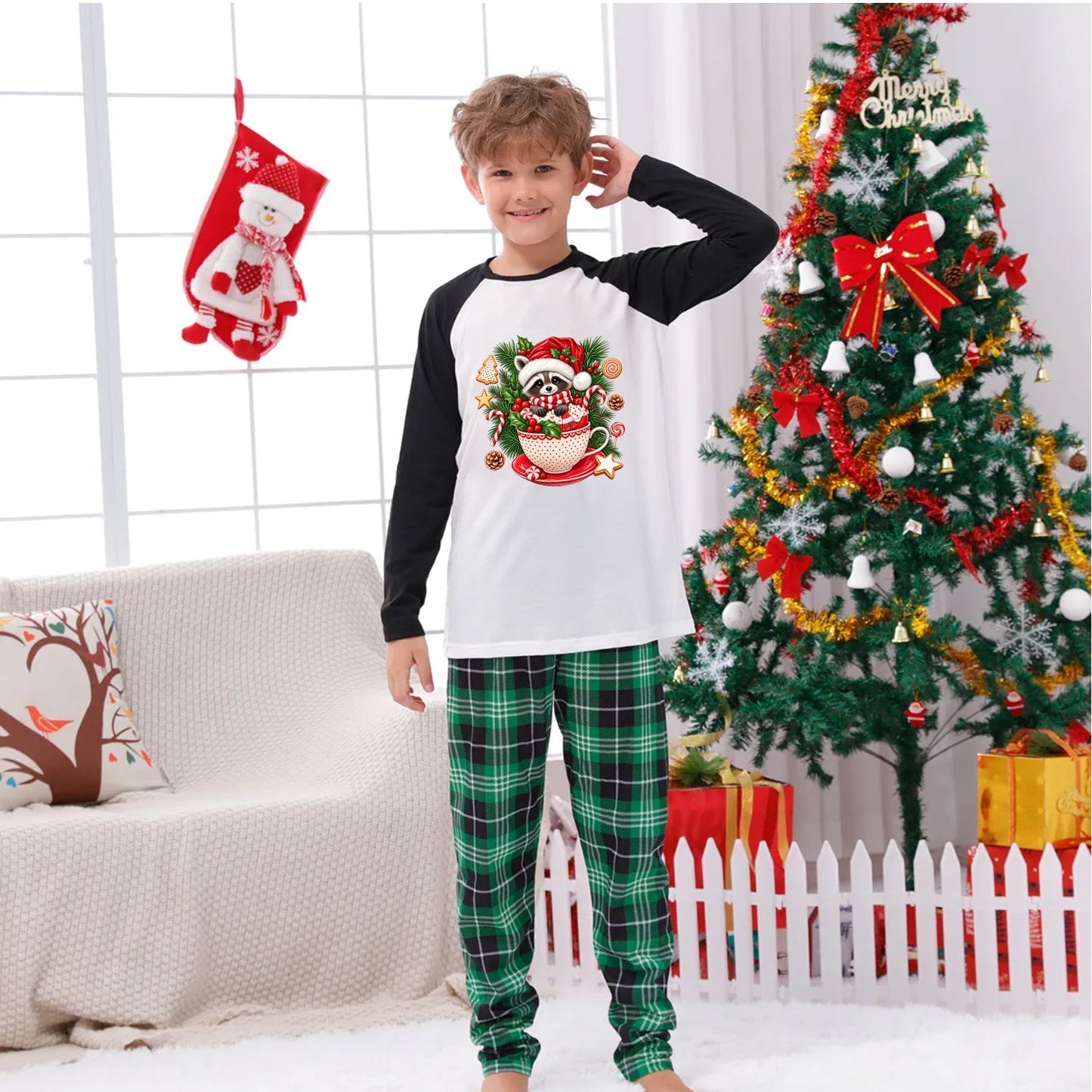 A young boy wearing a Christmas-themed raglan long-sleeve shirt and green plaid pajama pants stands in a decorated living room with a Christmas tree, stockings, and a couch.