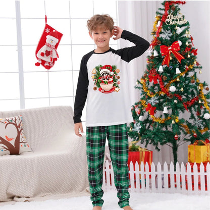 A young boy wearing a Christmas-themed raglan long-sleeve shirt and green plaid pajama pants stands in a decorated living room with a Christmas tree, stockings, and a couch.