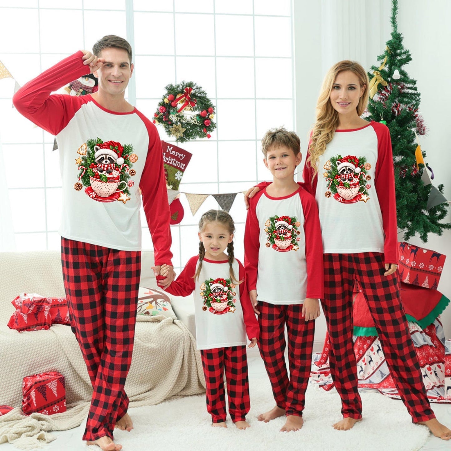 A family of four wearing matching Christmas-themed long-sleeve pajamas stands together in a decorated living room. The pajamas have red and black buffalo plaid legs with white fronts and red sleeves, featuring a festive Raccoon graphic.